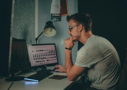 Focused man using a desktop computer in a dimly lit workspace with modern desk setup.